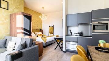 A modern hotel room with a bed, sofa, dining table, and kitchenette.