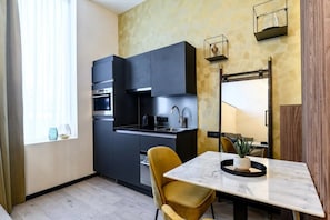 Una cocina compacta con muebles oscuros, horno empotrado y un pequeño comedor con mesa de mármol y una sola silla amarilla.