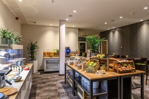 Ein Frühstücksbereich in einem Hotel mit einer Theke, auf der verschiedene Speisen, eine Kaffeemaschine und eine Sitzgelegenheit mit Tischen und Stühlen präsentiert werden.