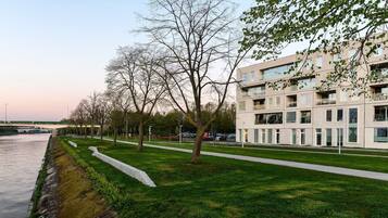 A modern building with a flat roof and large windows, situated next to a river with a paved walkway and trees.