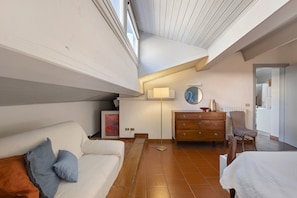 Ein Zimmer mit hohen Decken, einem weißen Sofa mit Kissen, einer Holzkommode, einer Stehlampe und Blick in den angrenzenden Raum durch eine offene Tür.