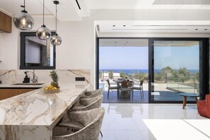 A modern kitchen with a marble countertop, hanging pendant lights, and a view of the ocean through large sliding doors. - Panoramic Sea View Villa in Hersonissos (Hersonissos)