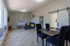 A modern living room with a glass dining table, a sofa with a yellow throw, and a large abstract painting on the wall. - Newly Renovated Quiet 2BR | WiFi, Parking & Heat Included—Sleeps up to 6! (London)