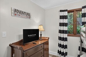 Een kamer met een flatscreen televisie op een houten dressoir, een lamp, een bord aan de muur en een raam met zwart-wit gestreepte gordijnen.