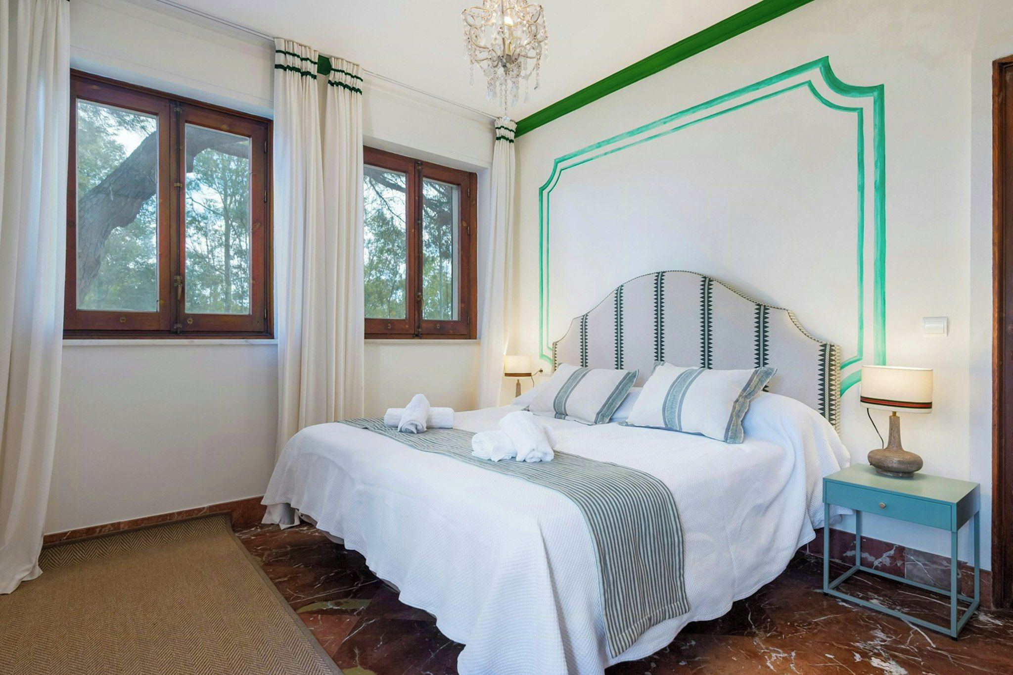 A hotel room with a bed, a nightstand, a lamp, and a view of trees through the window.