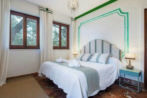 Ein Hotelzimmer mit Bett, Nachttisch, Lampe und Blick auf Bäume durch das Fenster.