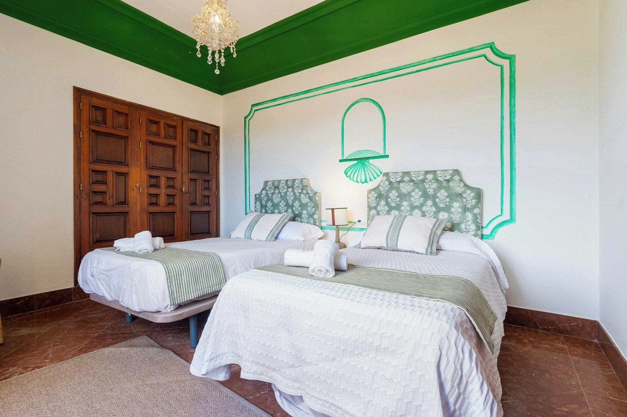 A hotel room with two beds, a chandelier, a green ceiling, and a wall with a decorative light fixture.