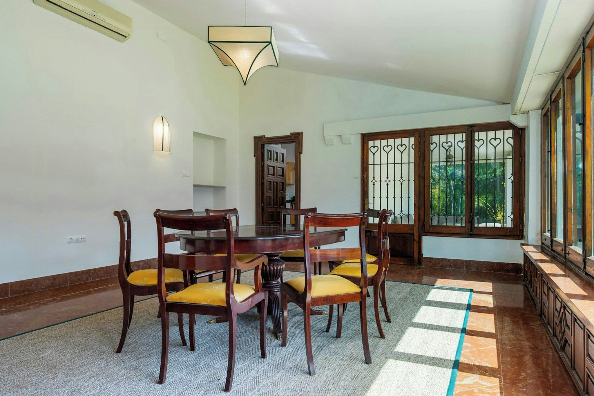 A dining area with a wooden table and chairs, a light fixture above, and a view through a window with decorative bars.