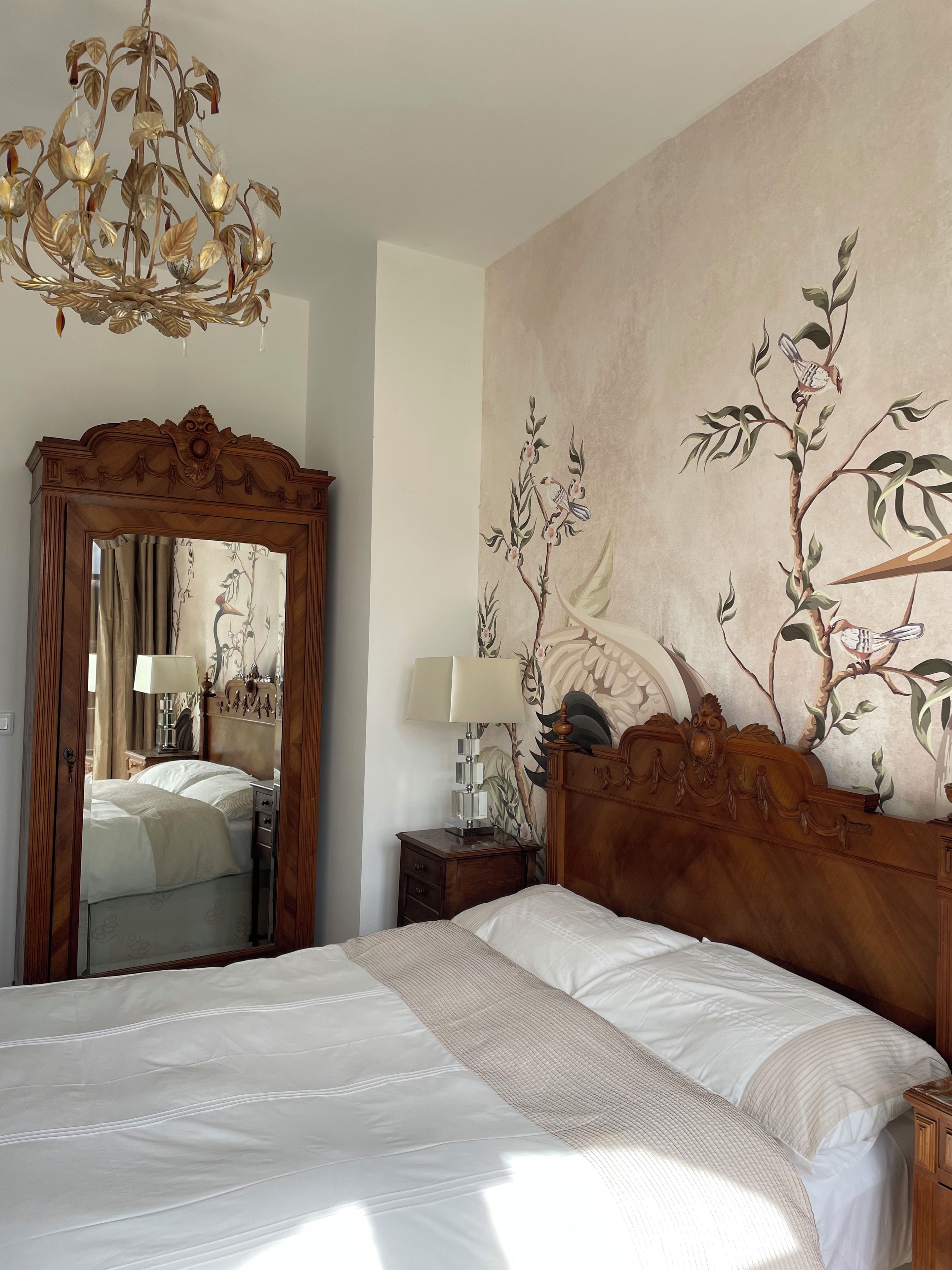 A bedroom with a large bed, a wooden headboard, a mirror, a nightstand, a lamp, and a wall mural.