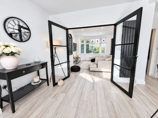 Ein modernes Wohnzimmer mit einer schwarz gerahmten Glastür zu einem angrenzenden Raum, einem weißen Sofa und einer an der Wand befestigten Uhr.
