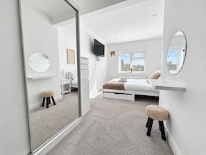 Ein modernes Schlafzimmer mit einem Bett, einem Fernseher, einem Fenster mit Jalousien, einem Spiegel und einem kleinen Hocker.
