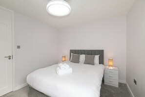 Ein ordentlich bezogenes Bett mit weißen Leinen und zwei aufgerollten Handtüchern, ein Nachttisch mit Lampe sowie eine Tür mit Griff.