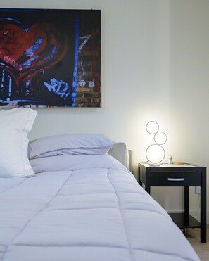Una cama bien tendida con ropa blanca, una mesita de noche negra con una lámpara y un cuadro abstracto grande en la pared.
