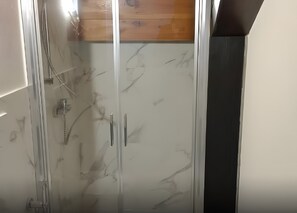 A bathroom with a glass shower enclosure, a wooden panel above the shower, and a white tiled wall.