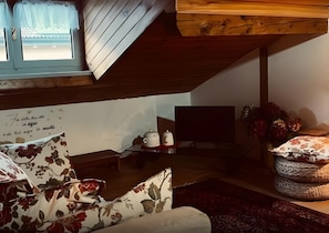 Kodikas olohuone, jossa on kalteva katto, kukkapainokuosilla verhoiltu sohva, pieni pöytä teekannuineen ja televisio.