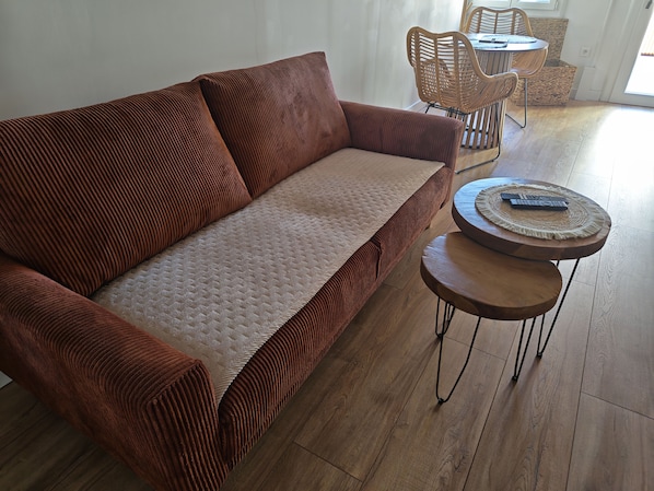 A brown sofa with a quilted cover, a round wooden coffee table with a remote control, and wicker chairs in a room with wooden flooring.