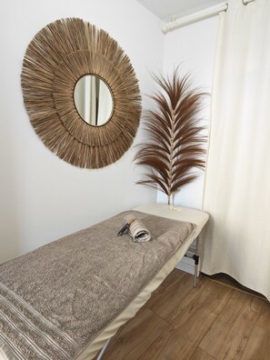 A room with a wooden floor, a bed with a textured blanket, a round mirror, and a decorative plant.