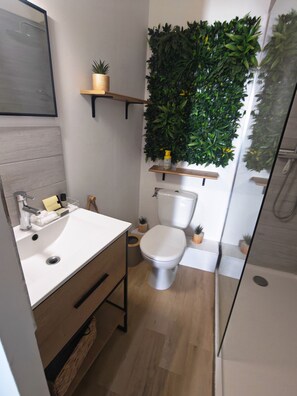 Un baño moderno con lavamanos blanco, inodoro y zona de ducha. Hay estantes con plantas y artículos de aseo.