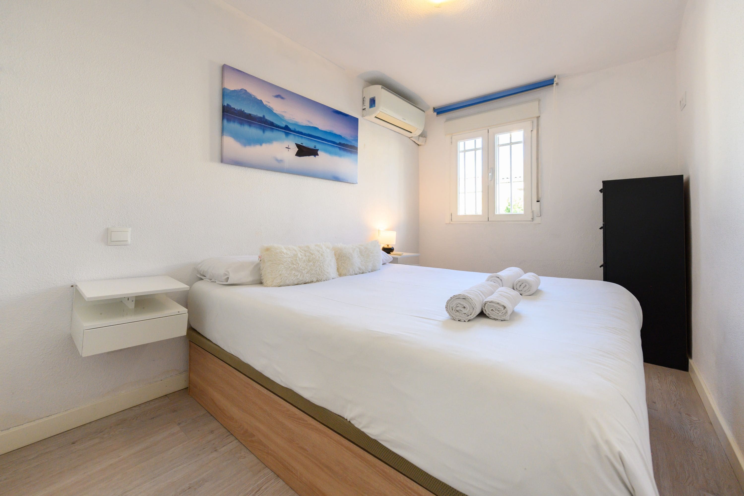 A bedroom with a bed, a nightstand, a wall-mounted air conditioner, a window, and a painting of a landscape.