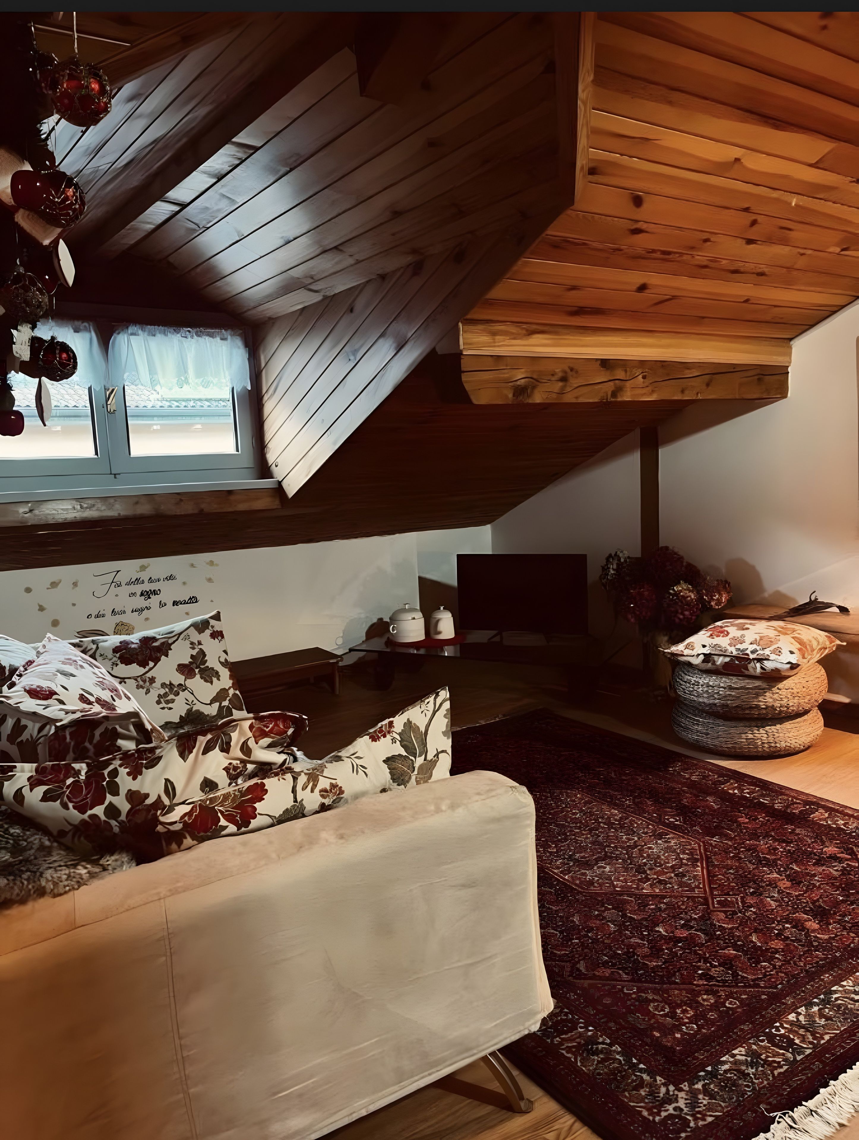 Ett mysigt sovrum på loftvåningen med ett sluttande tak, en säng med blommiga kuddar, en mörk, mönstrad matta och ett litet bord med dekorativa föremål.