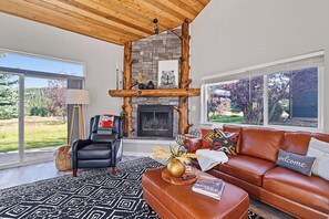 A modern living room with a stone fireplace, leather furniture, and a large window overlooking a garden. - Summit & Stream at Candlelight Meadows (Big Sky)