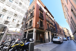 Una calle estrecha con motocicletas estacionadas y un coche blanco. Se ve un edificio con fachada roja y balcones.