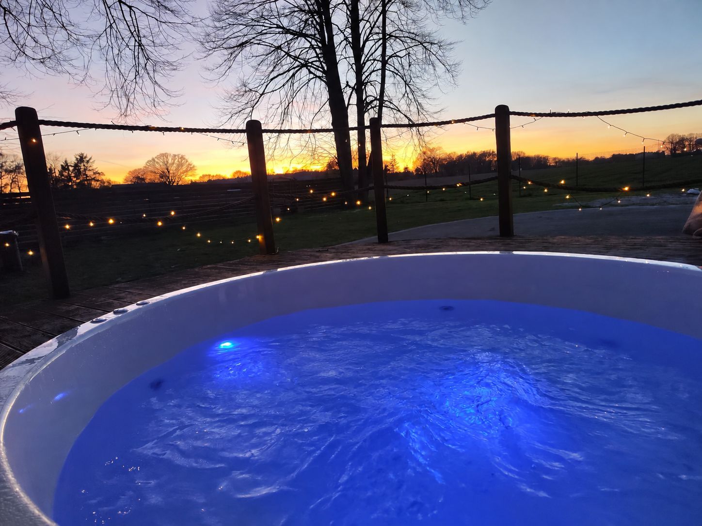 A hot tub with a blue light, surrounded by a fence and string lights, with a sunset in the background.