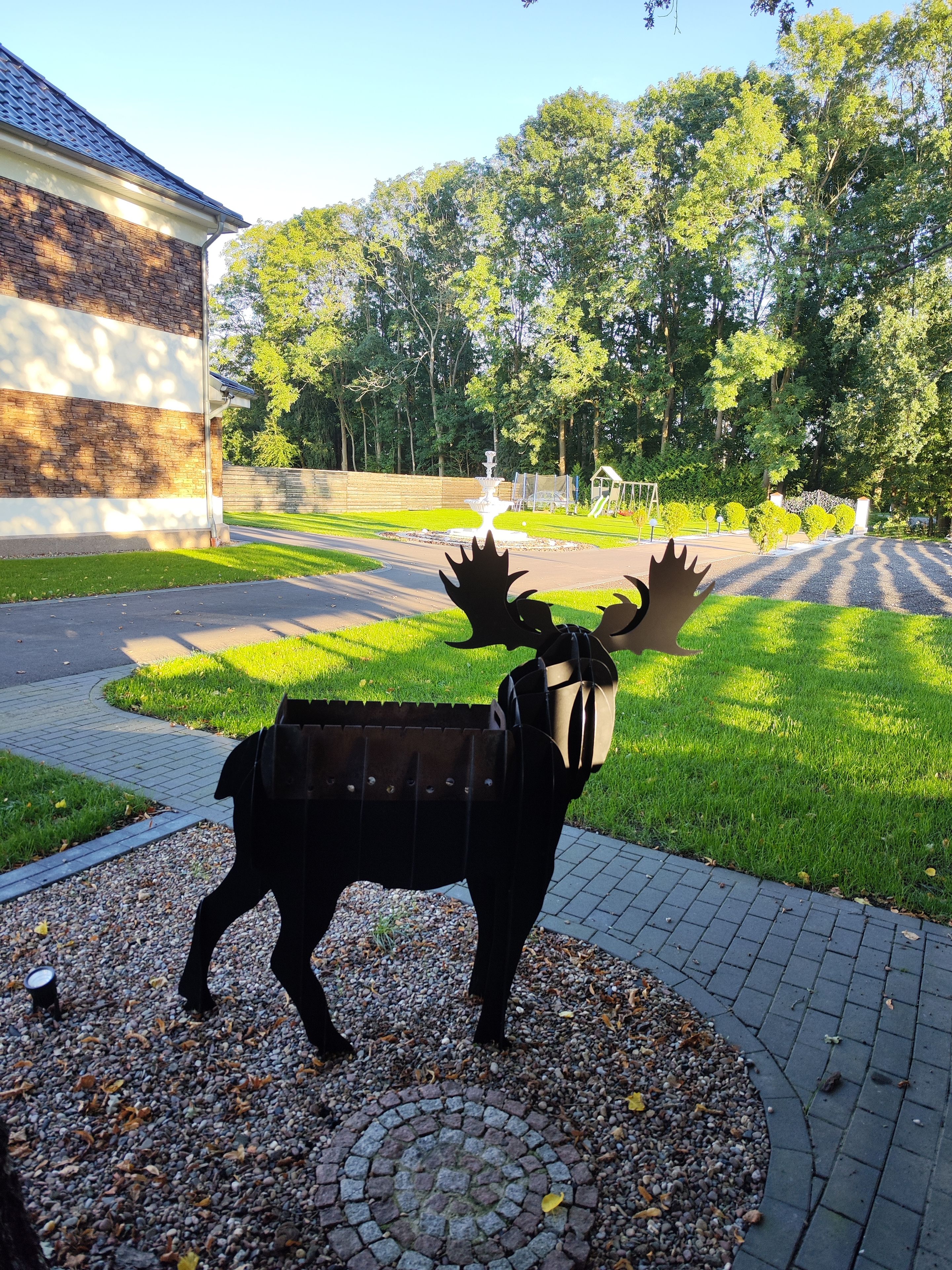A sculpture of a moose in a park setting with a paved walkway, trees, and a building in the background.