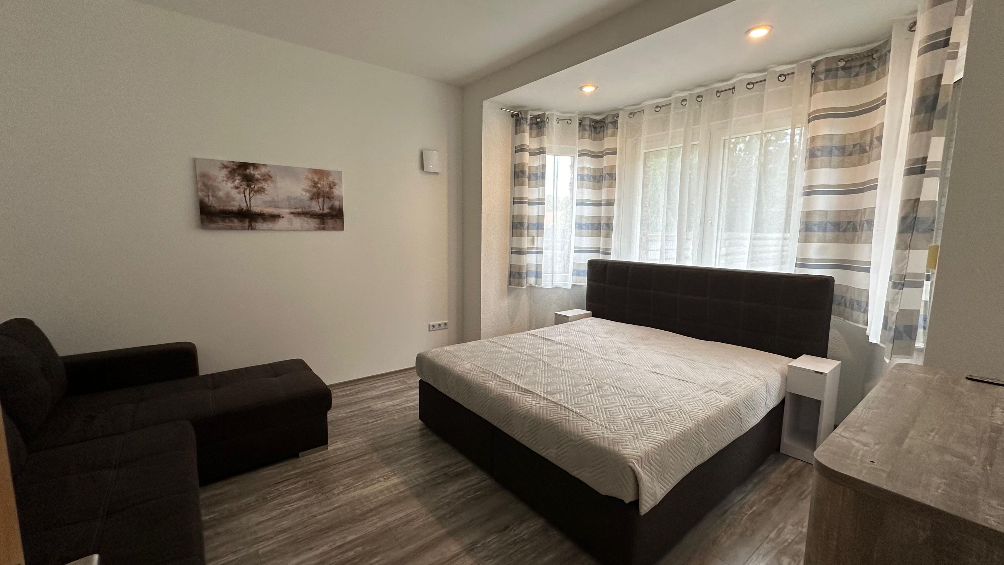 A bedroom with a bed, a chair, a bedside table, a painting on the wall, and a window with curtains.