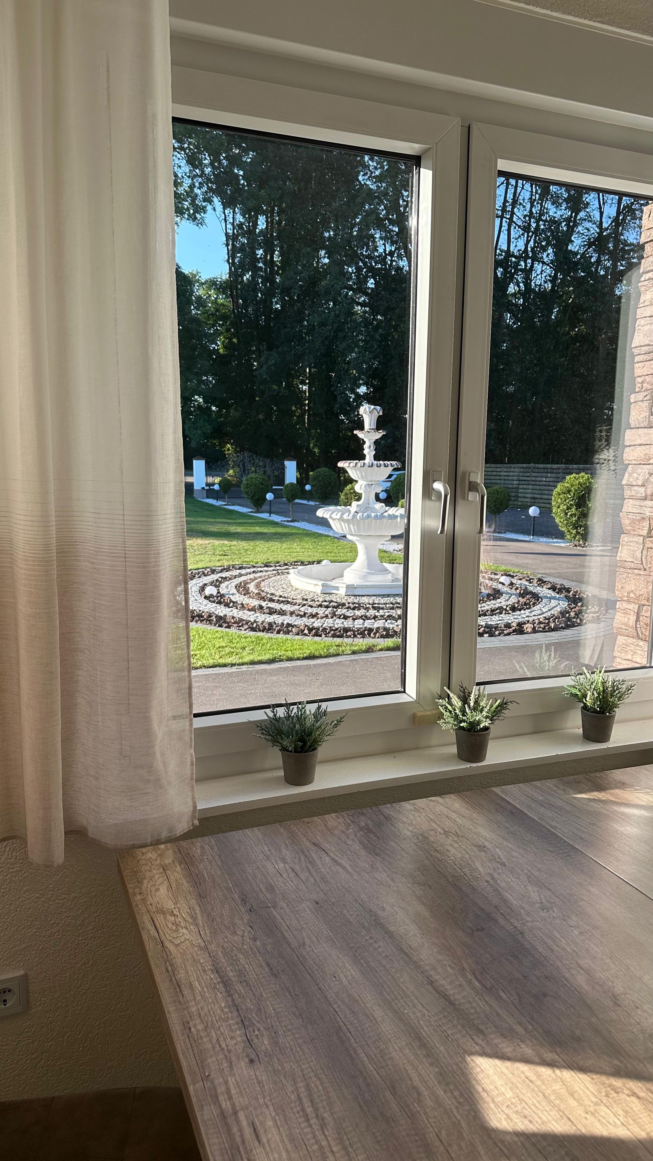 Ett rum med ett stort fönster med utsikt över en trädgård med en fontän och krukväxter.