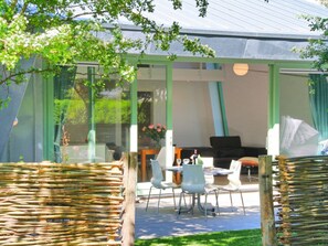 An outdoor seating area with chairs and a table, a glass enclosure, and a view of a building with a green roof. - Vakantievilla Sint Maartenszee (Sint Maartensvlotbrug)
