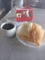Uma mesa com uma xícara de café branco, um prato de pastéis fritos e uma caixa vermelha com o logo de um coração.
