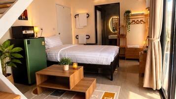 A modern hotel room with a bed, a desk, a chair, a small table, a plant, a mirror, a window with curtains, and a staircase.