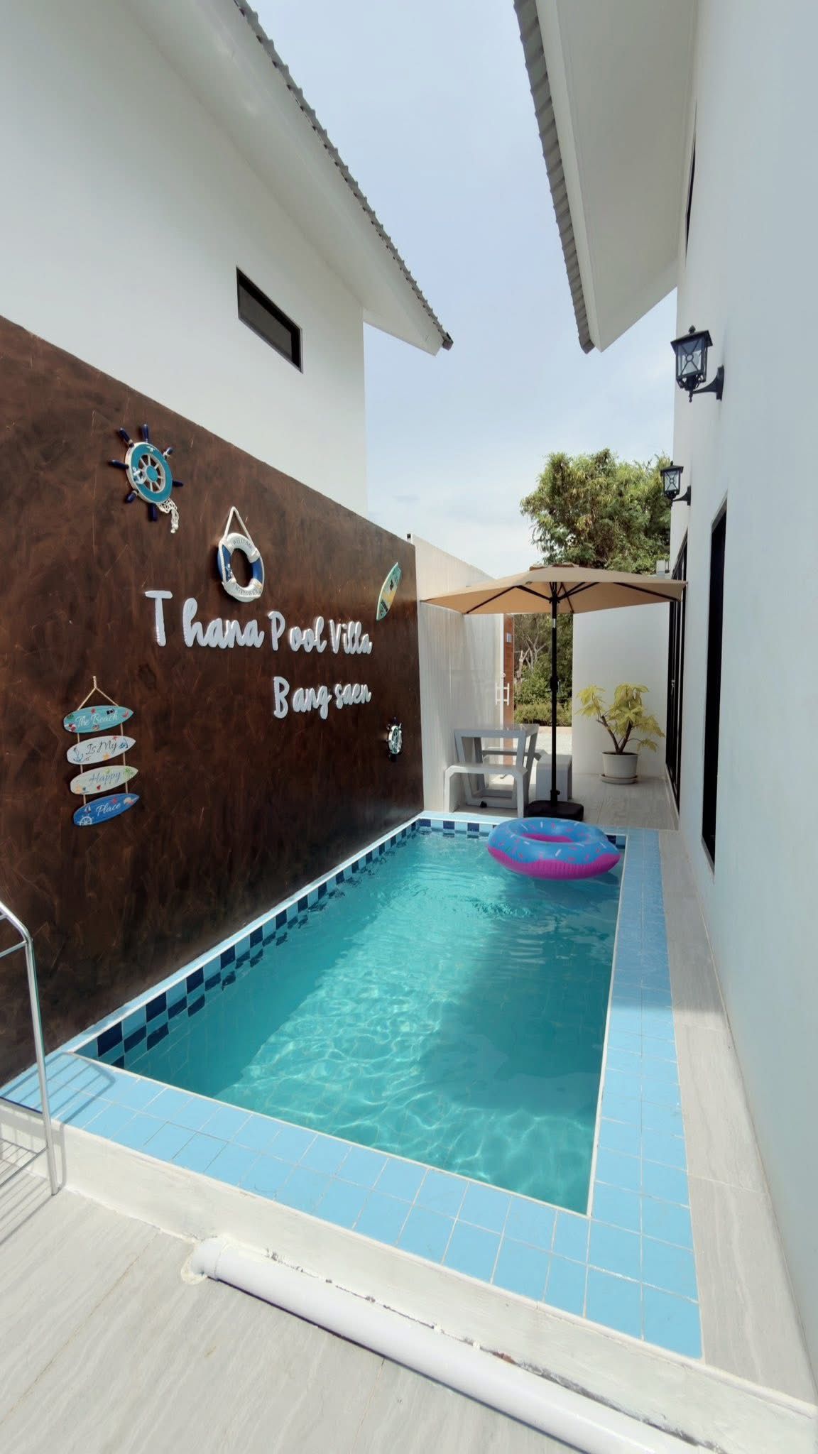 Una piscina con borde de azulejos azules y blancos, una silla de descanso y una planta en maceta.