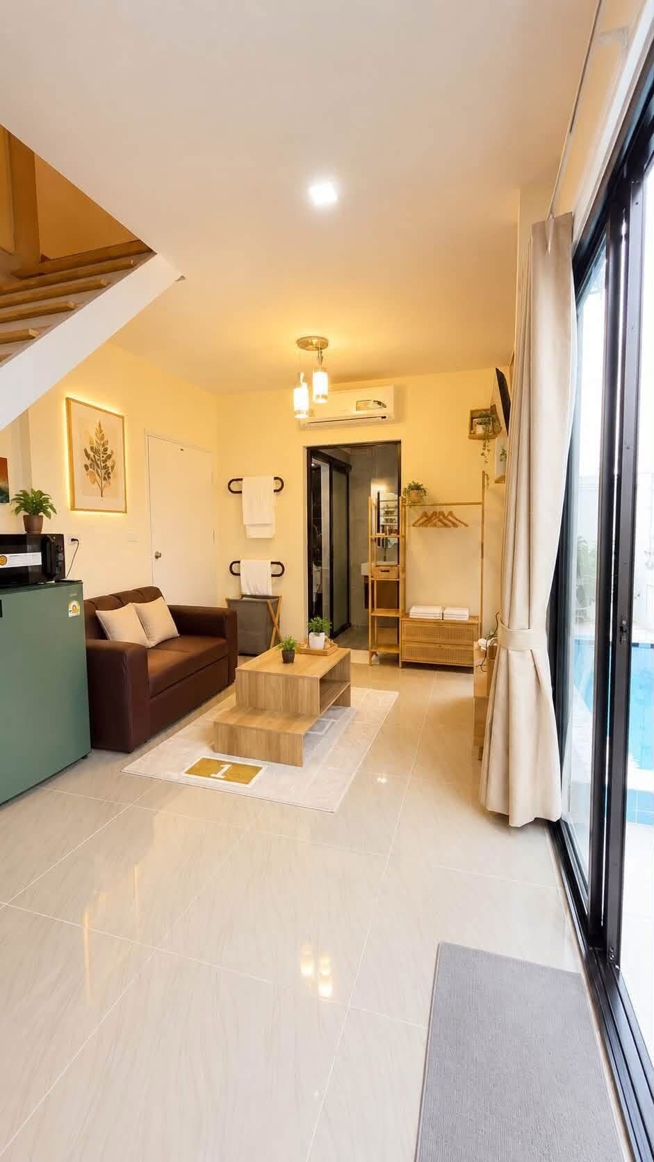 A modern living room with a sofa, coffee table, and a staircase. There is a door leading to another room and a window with curtains.