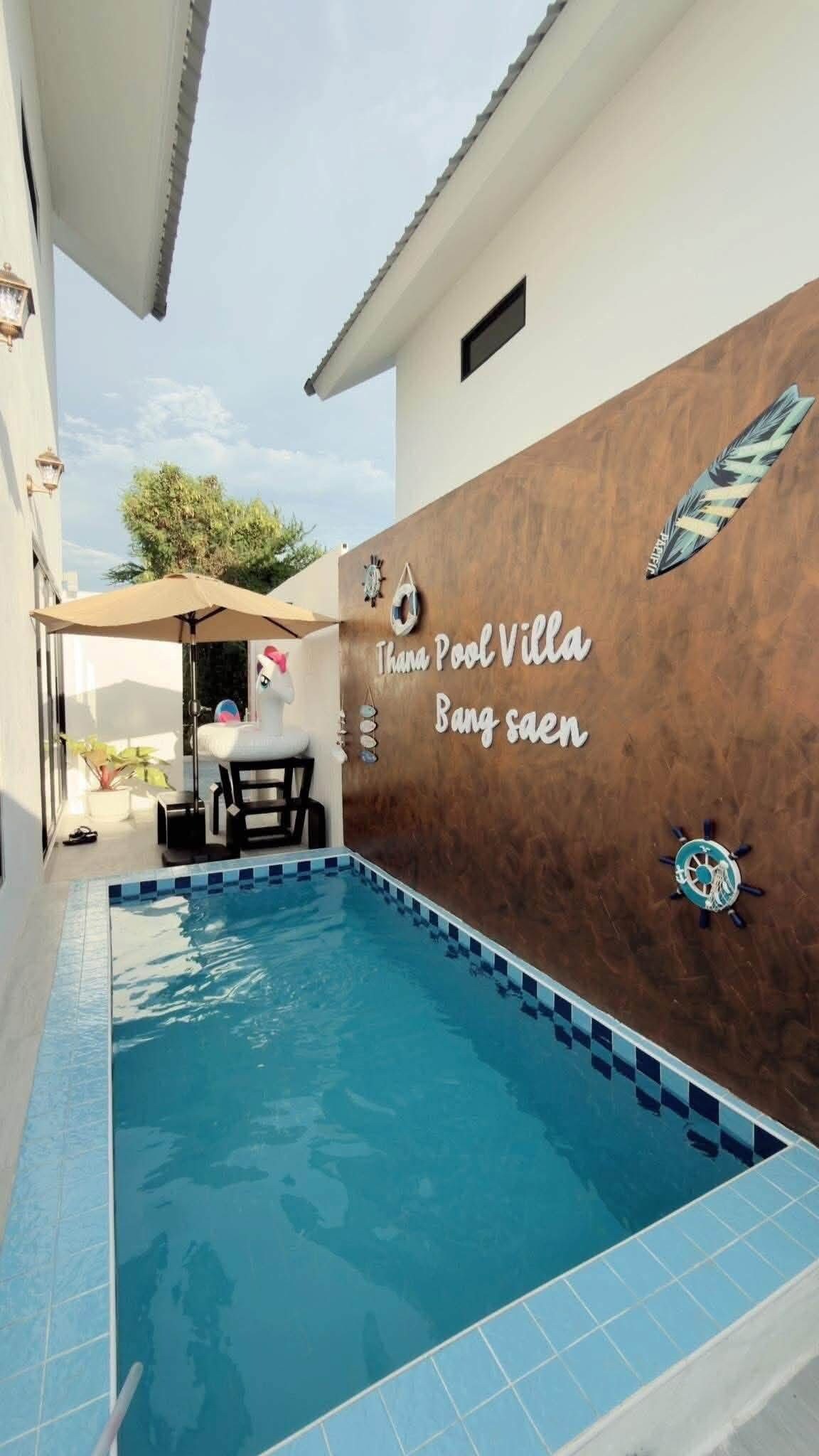 A swimming pool with a tiled edge, a wall with decorative elements, and a sign that reads 'Thana Pool Villa Bang Saen.'