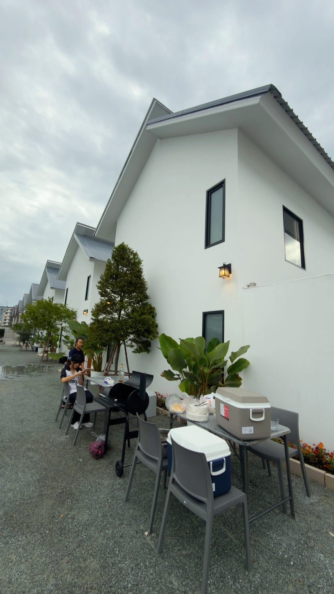 A row of white buildings with a flat roof, outdoor seating area with chairs and tables, and a large potted plant.