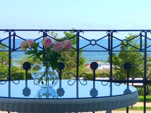 A round table with a clear glass vase containing flowers, set against a metal railing overlooking a beach. - Villa Fortuna BY THE Sea, Apartment 5 (Heringsdorf)