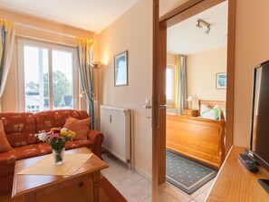 Ein Hotelzimmer mit Sofa, einem Tisch mit Blumenvase, einem Holztisch mit Fernseher und Fernbedienung sowie Blick auf das angrenzende Zimmer.
