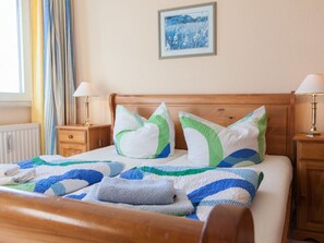 Ein ordentlich bezogenes Bett mit gestreifter Decke und Kissen, ein Nachttisch mit Lampe, ein gerahmtes Bild an der Wand und ein Fenster mit Vorhängen.