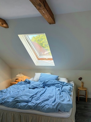 Ein Schlafzimmer mit Oberlicht, einem Bett mit gestreifter Bettwäsche, einem Nachttisch mit Lampe und einem kleinen hölzernen Hocker mit einer Vase.
