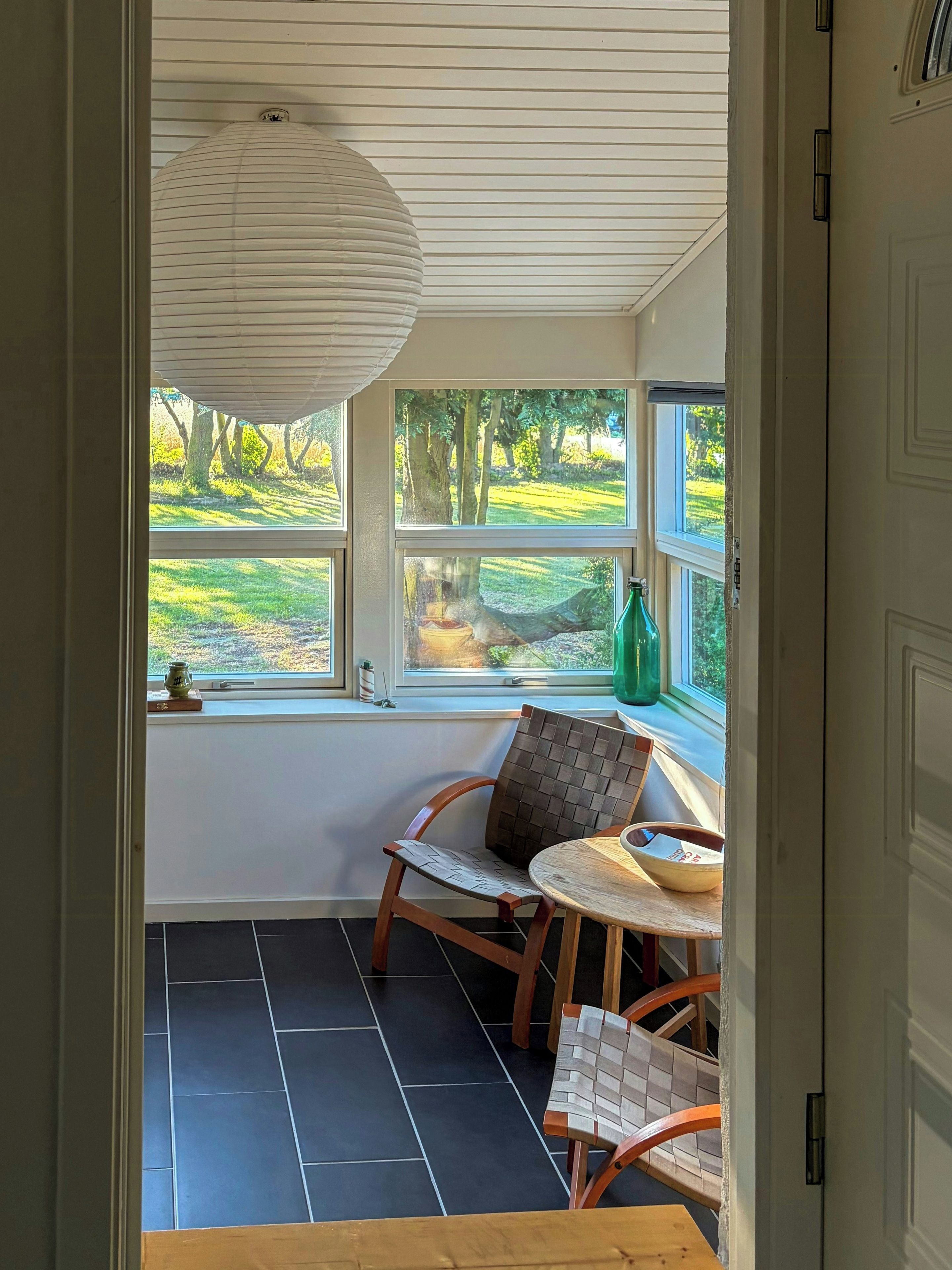 Une pièce donnant sur un champ verdoyant grâce à de grandes fenêtres, avec une chaise en bois, une petite table et une bouteille verte posée sur le rebord de la fenêtre.