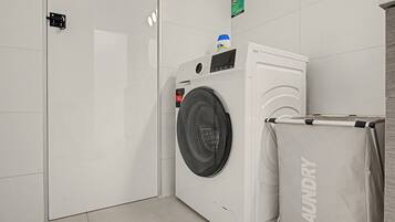 A laundry facility with a front-loading washing machine, a drying rack, and a trash bin.