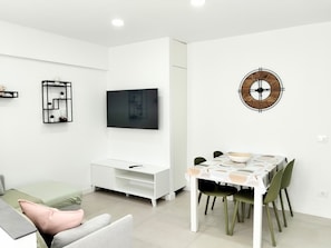 Una sala moderna con un televisor de pantalla plana, un mueble blanco para equipo multimedia, una mesa de comedor con sillas y un reloj de pared.