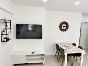 Una sala moderna con televisor de pantalla plana en la pared, mueble blanco para entretenimiento, una mesa de comedor con sillas y un reloj decorativo en la pared.