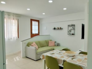 Una sala de estar moderna con un sofá, una mesa de comedor con sillas, un ventanal con cortinas y estantes en la pared con objetos decorativos.