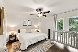 A bedroom with a bed, a ceiling fan, a nightstand, a lamp, a window with curtains, and a view of greenery outside. - Relaxing Pinehurst Getaway - 2 Miles to The Cradle & 5 Minutes to The Village (Pinehurst)