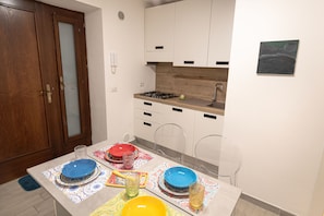 A compact kitchen with a wooden door, white cabinets, a gas stove, and a dining table set with colorful plates and glasses. - One Bedroom apartment in the centre of Assisi (Assisi)