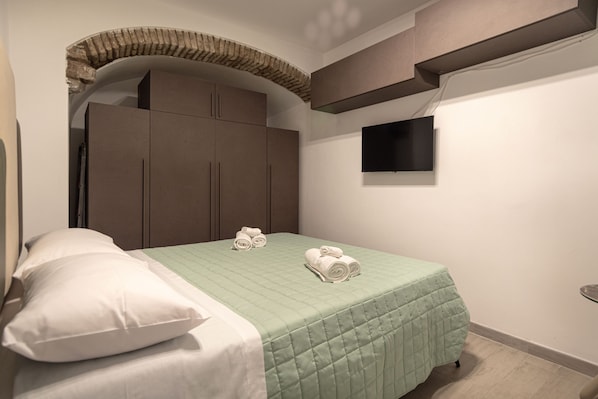 Een slaapkamer met een bed, kussens, een groene geborduurde spreideken, een flatscreen televisie aan de muur en een kledingkast.