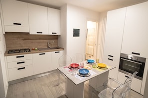 Een kleine keuken met witte kasten, een inbouwoven en een eethoek met transparante stoelen.